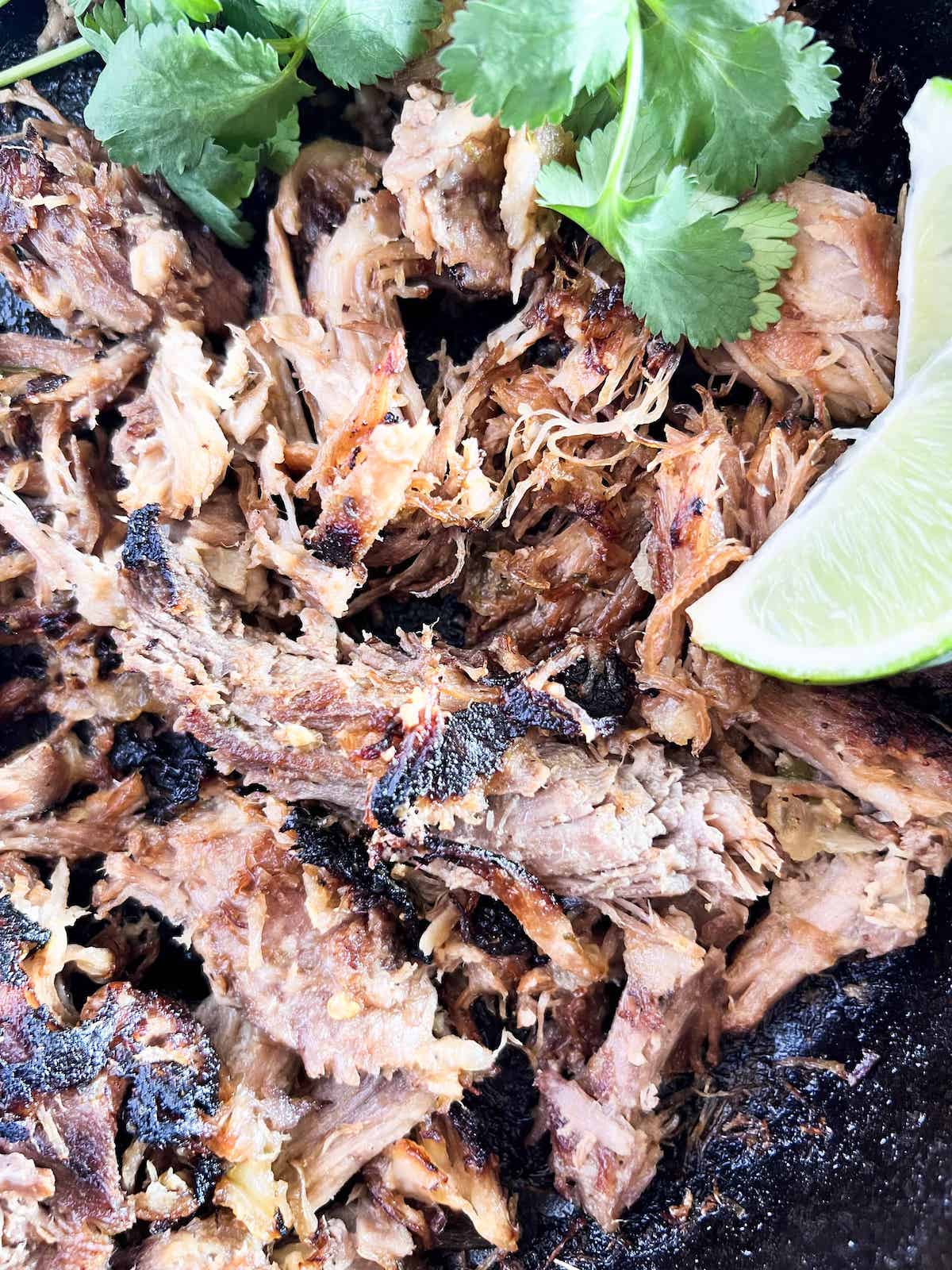 Close up of crispy pork carnitas in a cast iron skillet with cilantro and lime.