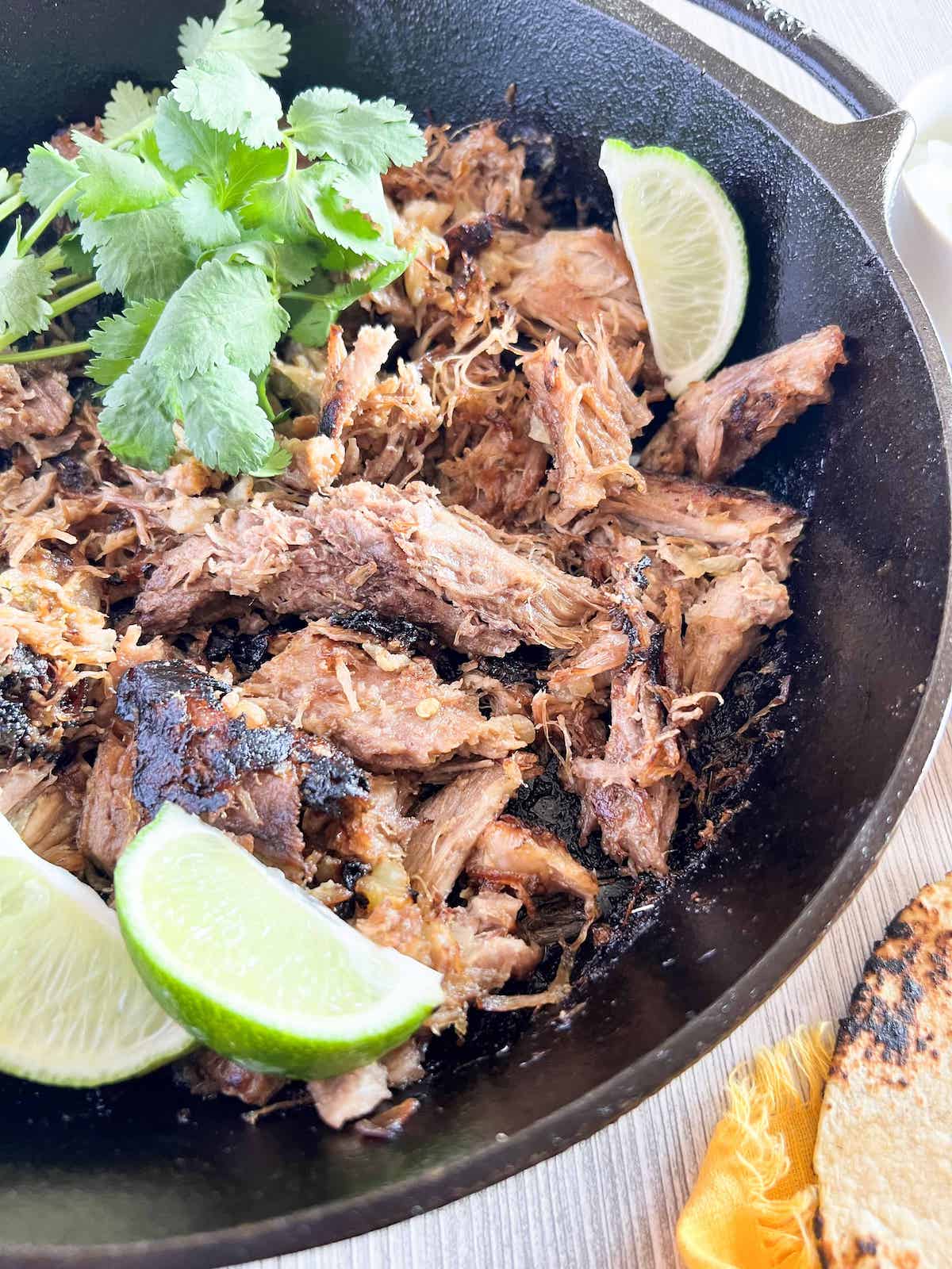 Cast iron skillet with crispy pork carnitas garnished with lime wedges and cilantro.