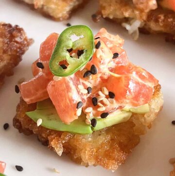 Close up of crispy rice cake topped with avocado, spicy tuna, sesame seeds and a slice of Serrano pepper on a white platter.