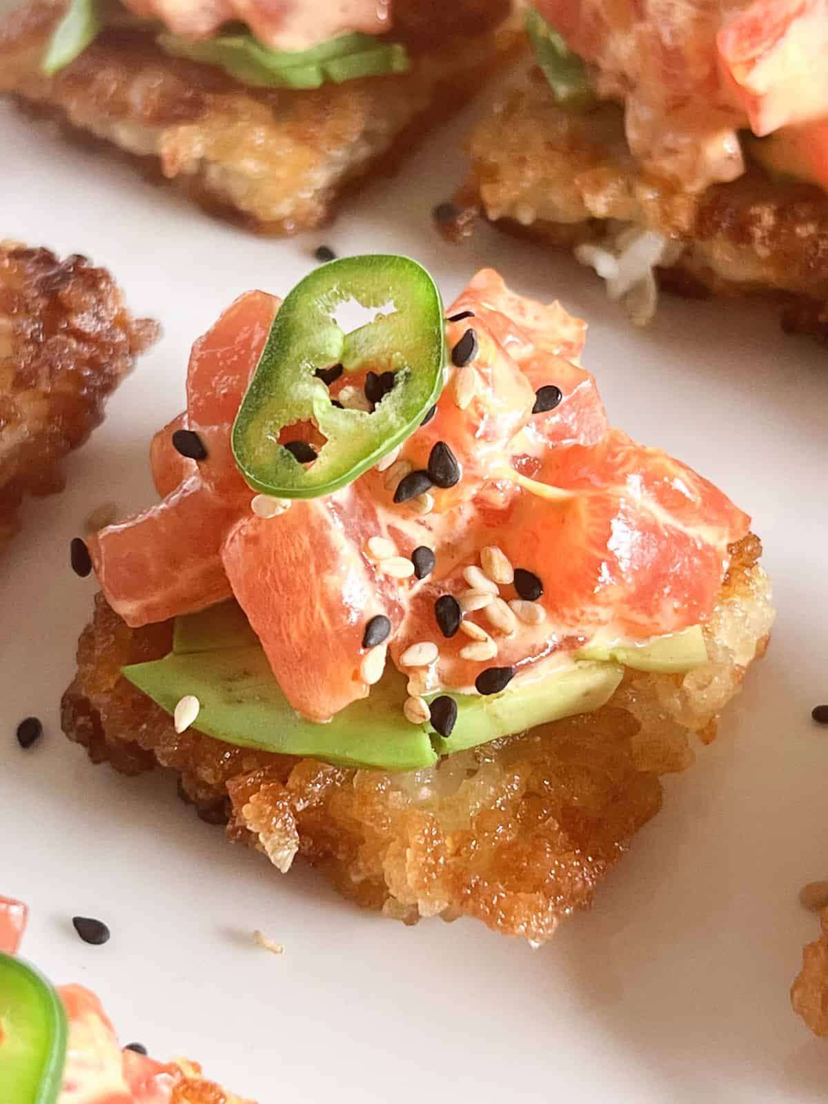 Close up of crispy rice cake topped with avocado, spicy tuna, sesame seeds and a slice of Serrano pepper on a white platter.