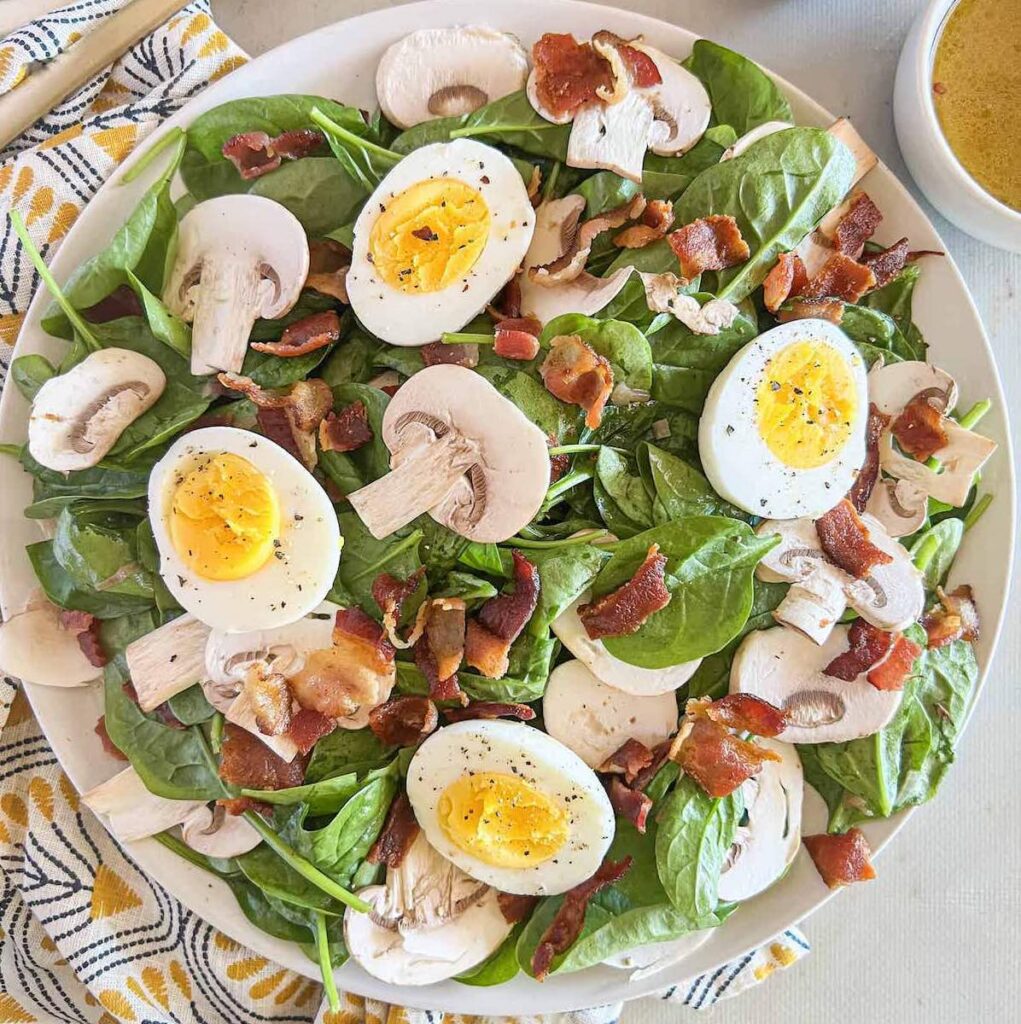 White plate of baby spinach salad topped with sliced mushrooms, chopped bacon and sliced hard boiled eggs.