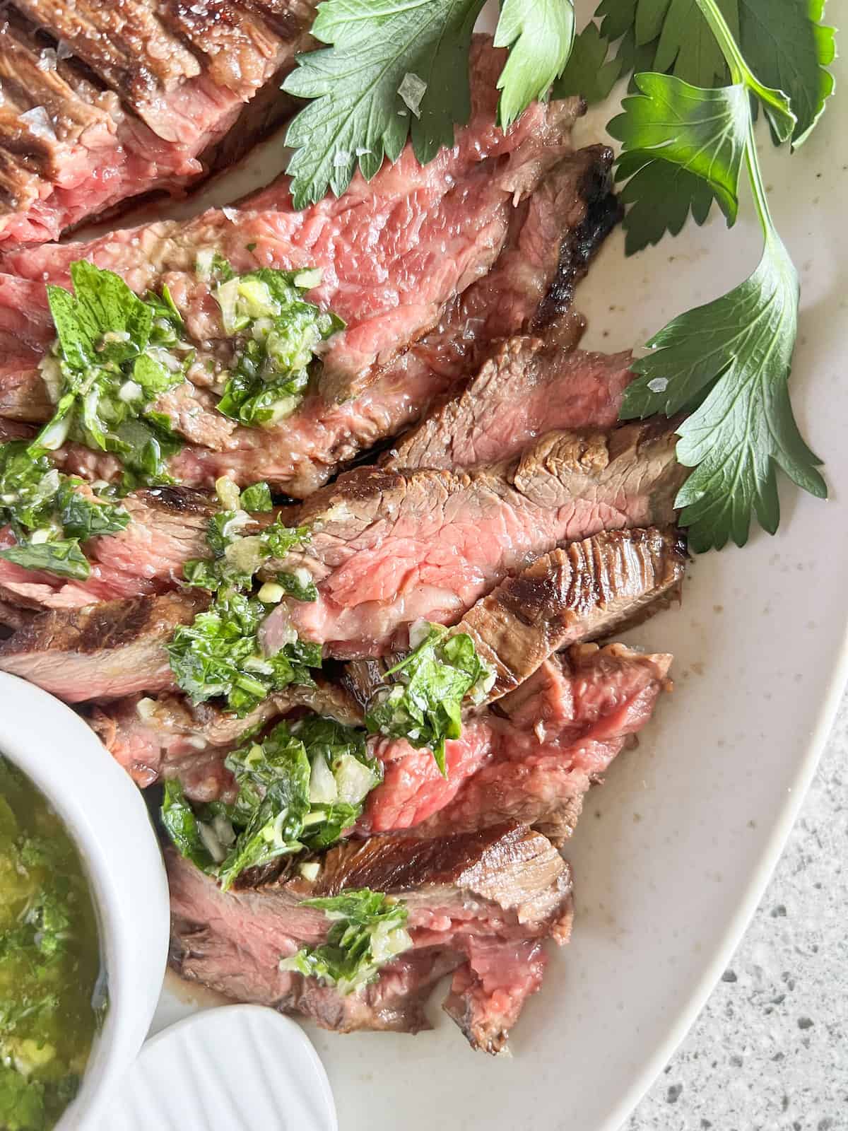 Close up of slices of medium rare bavette steak topped with chimichurri sauce on a white platter.