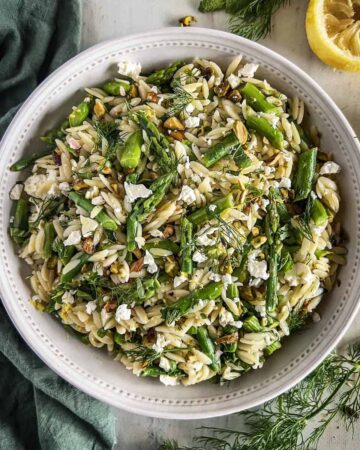 Orzo asparagus salad in a white bowl with a green napkin, herbs and squeezed lemon half in the background.