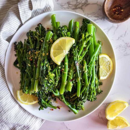 Sauteed broccolini garnished with lemon zest, red pepper flakes and lemon slices on a white plate.