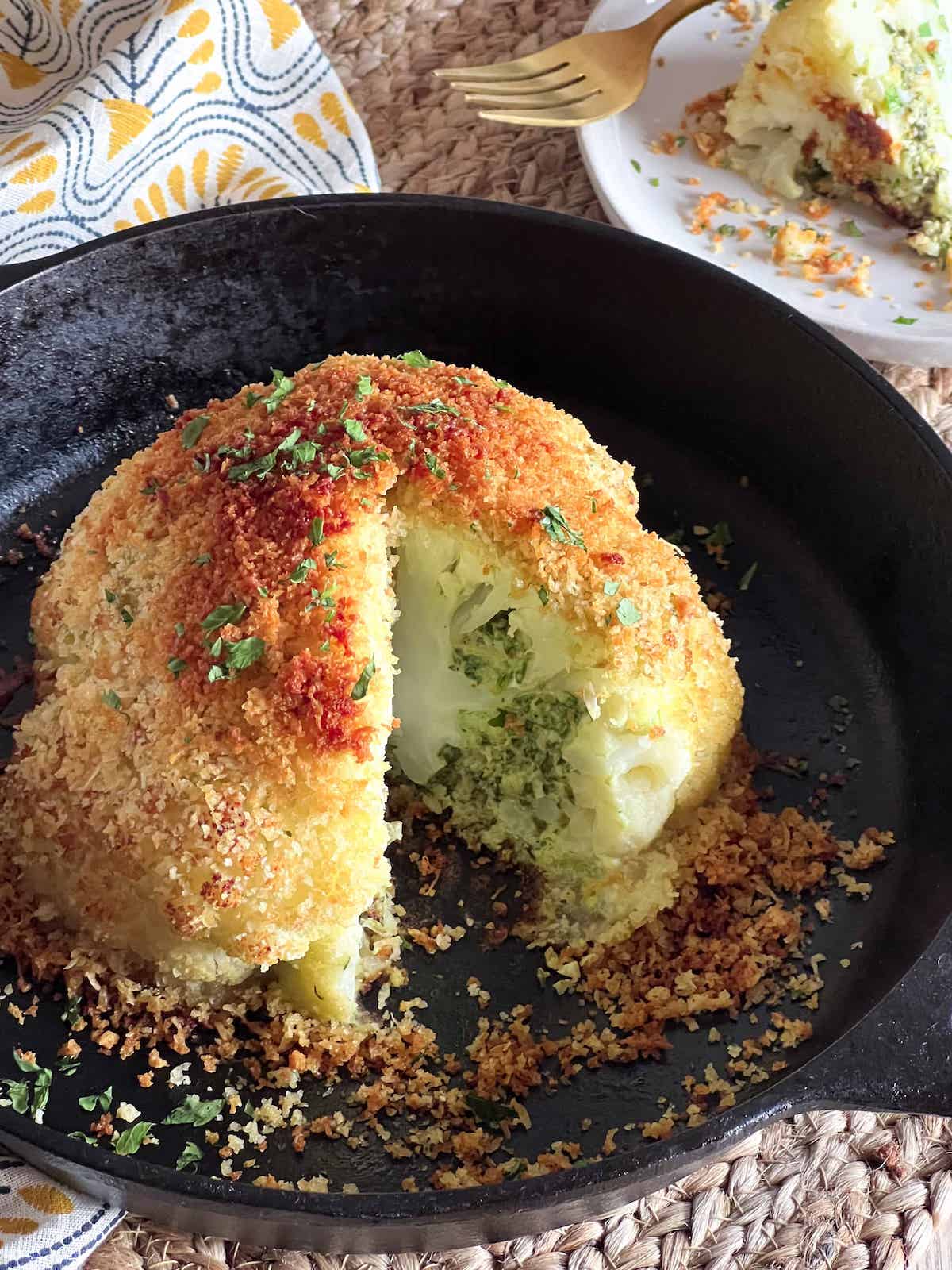 Whole breaded baked cauliflower in a cast iron skillet with a slice cut out.