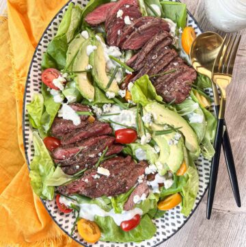 Grilled steak salad on an oval platter with a yellow napkin, gold silverware and a jar of blue cheese dressing.