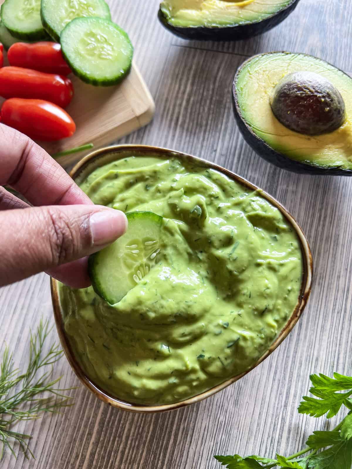 Brown hand dipping cucumber slice into small bowl of avocado ranch dressing.