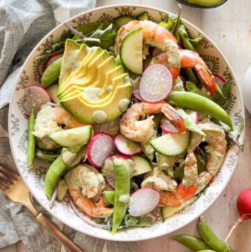 Baby kale salad topped with shrimp, sliced avocado, radish, cucumber and snap peas in a patterned bowl.