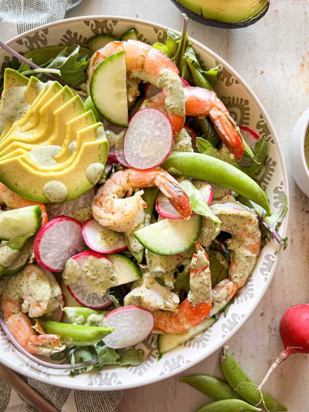 Close up of baby kale salad topped with shrimp, sliced avocado and veggies, drizzled with dressing.