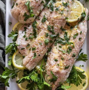 Baked walleye on a white platter with lemon slices and fresh herbs.