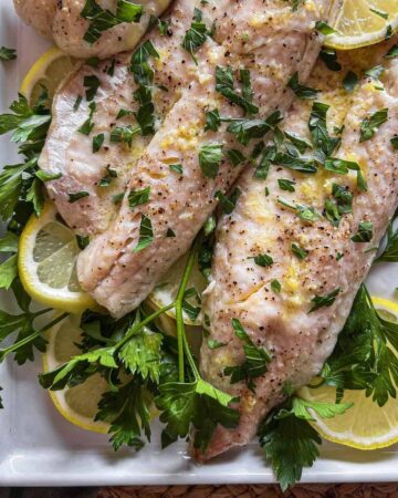Baked walleye on a white platter with lemon slices and fresh herbs.
