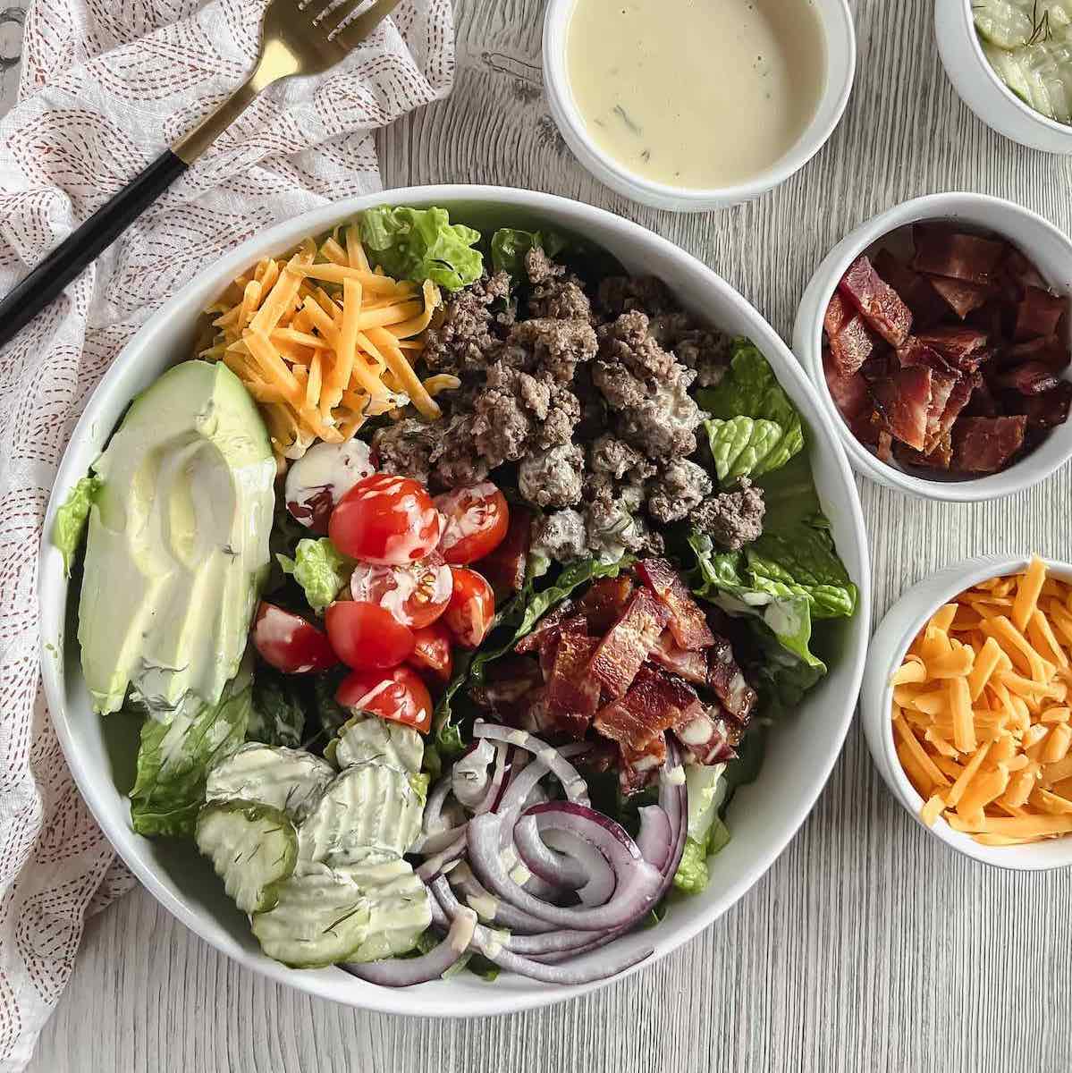 oaded burger bowl in a white dish, surrounded by smaller white bowls with burger toppings and a gold fork on a napkin.