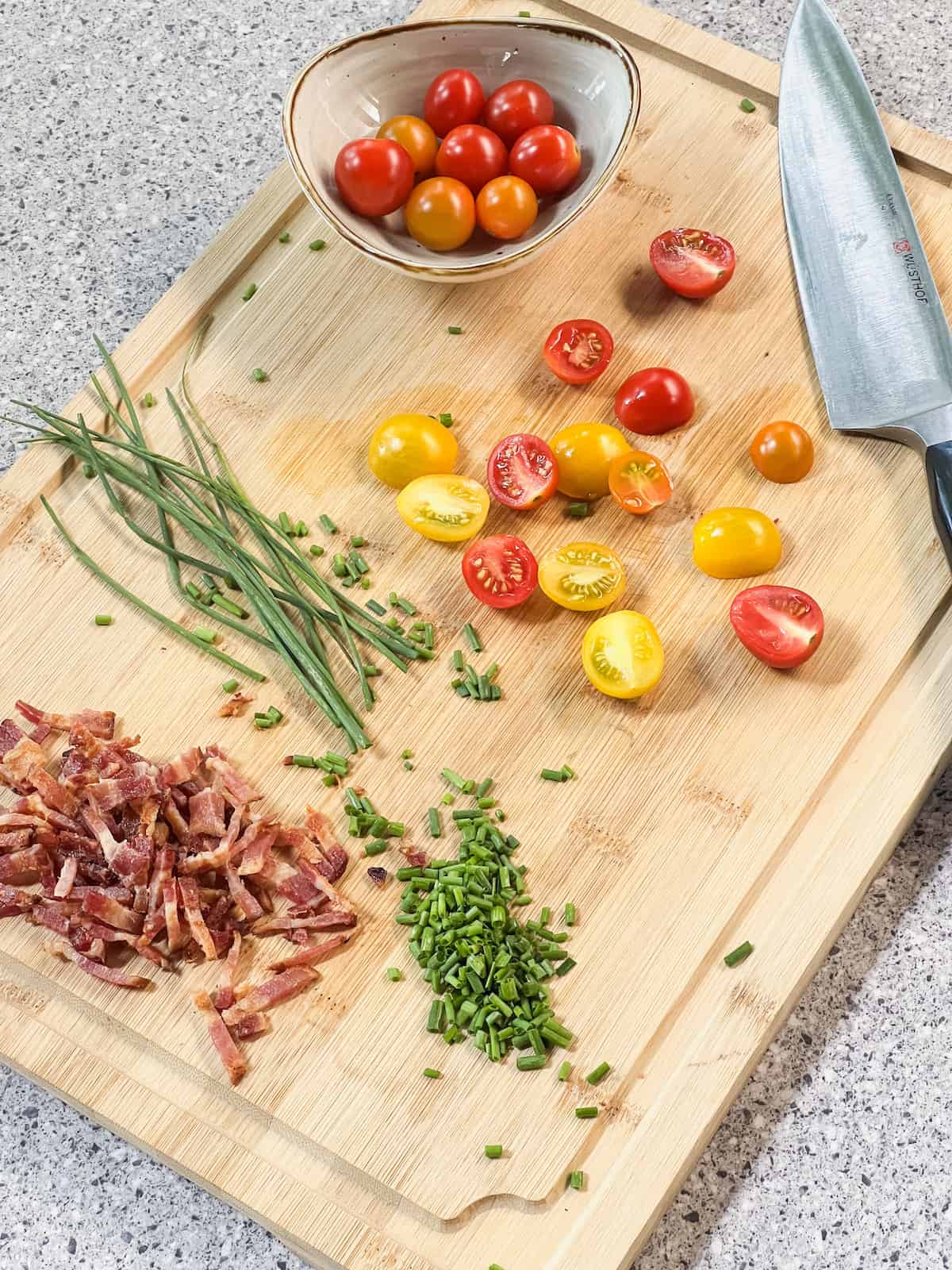Halved cherry tomatoes, chopped chives and bacon on a wood cutting board with a knife.