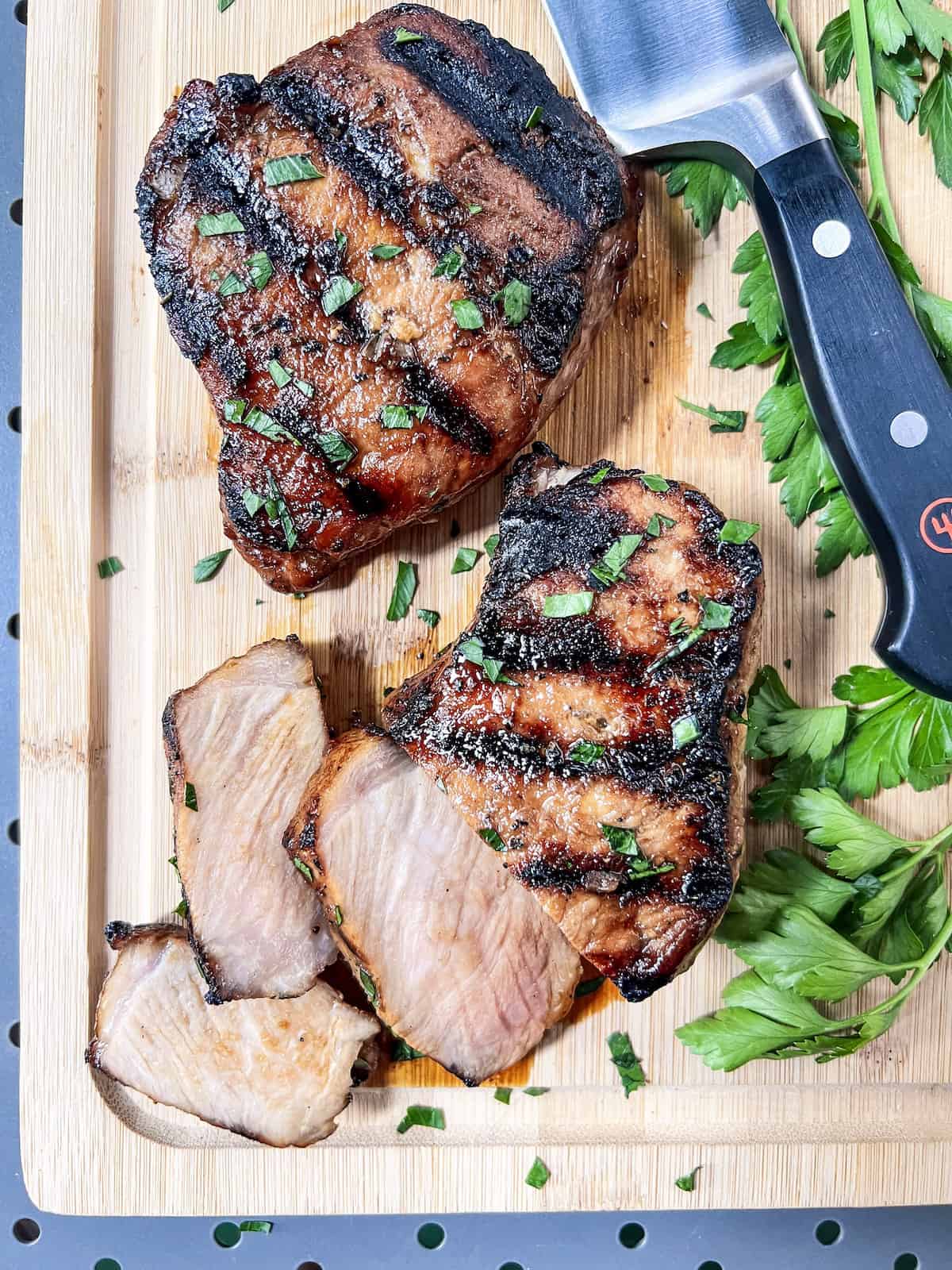 Grilled pork chops, garnished with parsley, on a wood cutting board with a knife. 