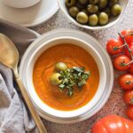 Bowl of roasted red pepper and tomato soup garnished with green olives and basil with a bowl of olives and fresh tomatoes in the background.