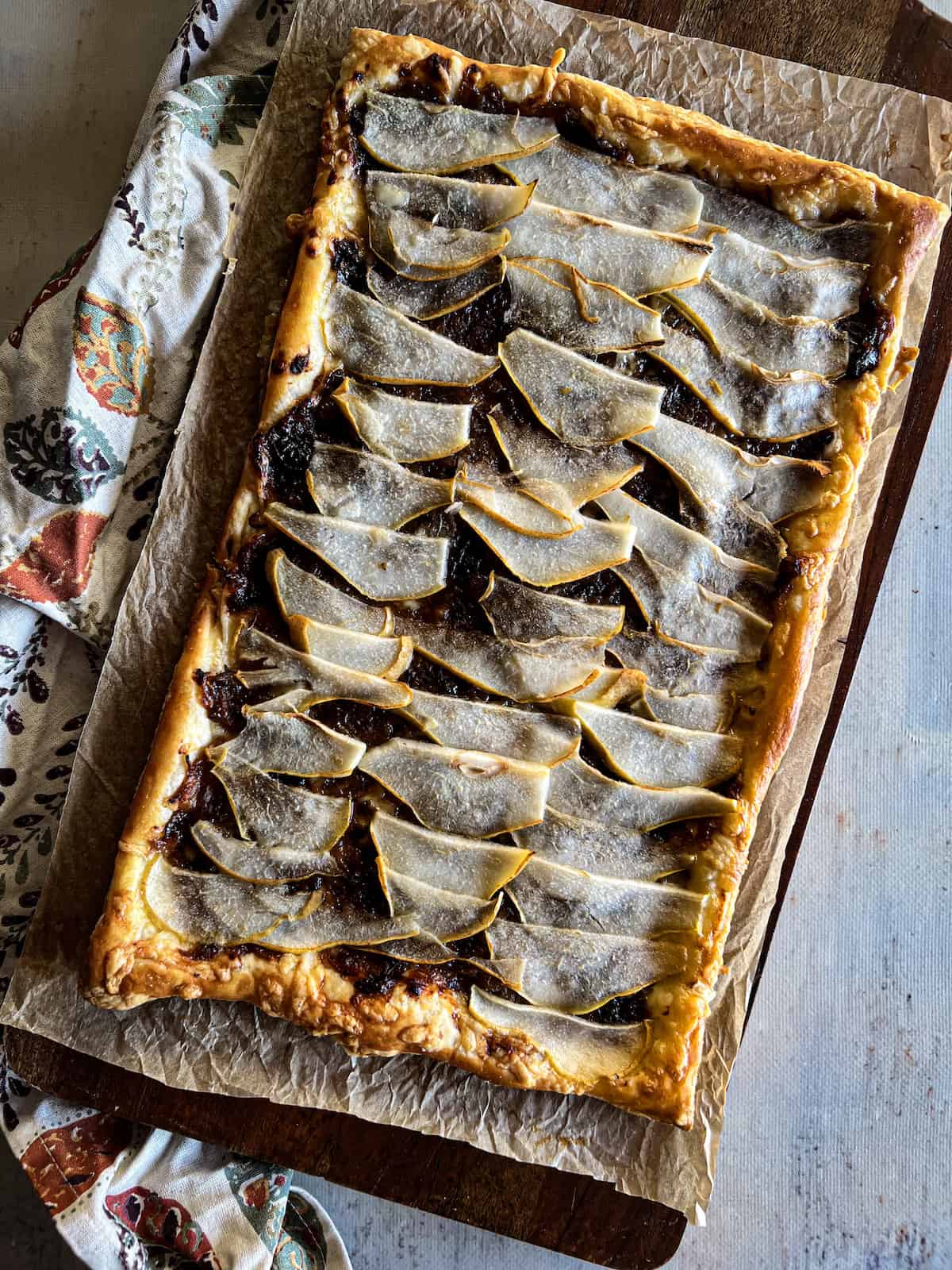 Pear, caramelized onion and prune tart on parchment paper on a wood board with a leaf patterned napkin.