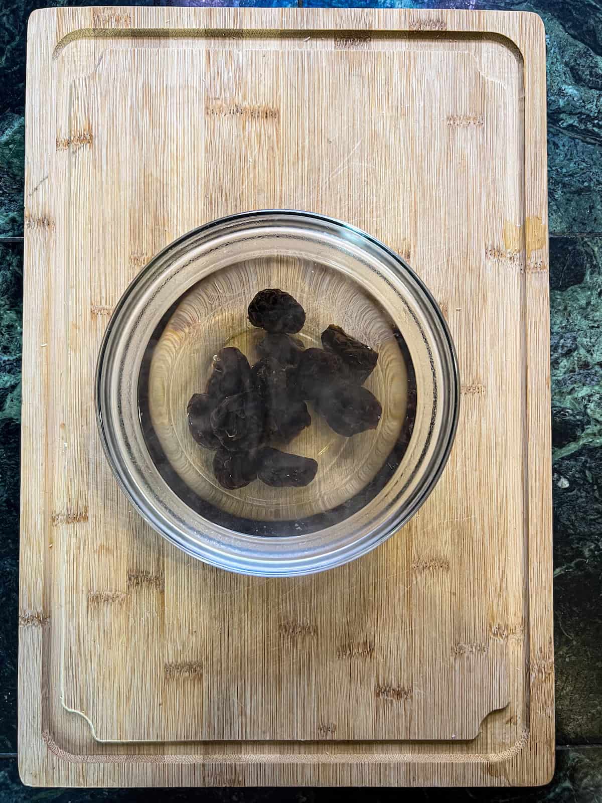 Prunes soaking in hot water in a glass bowl on a wood cutting board.