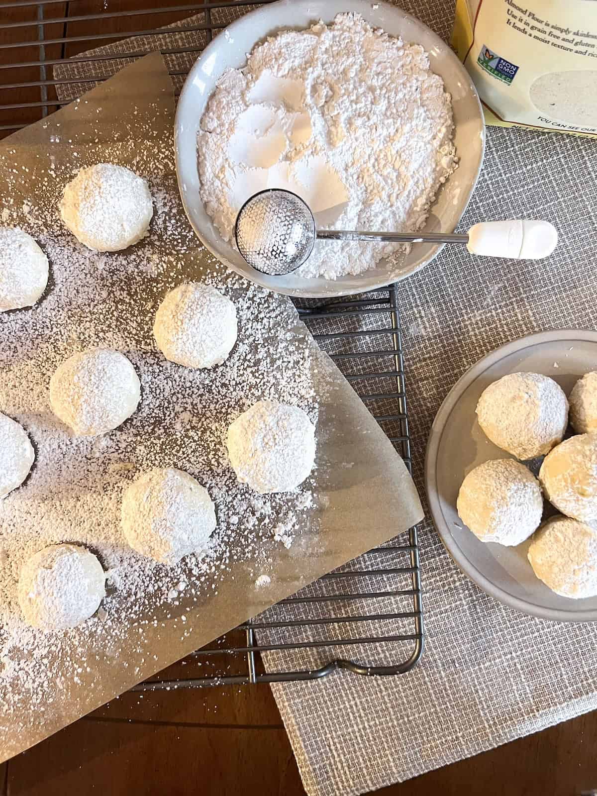 Snowball cookies on parchment paper next to a plate of powdered sugar with a bakers dusting wand.