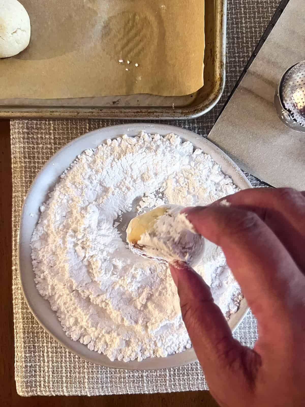 Brown hand rolling baked cookies in powdered sugar.