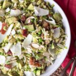 Shredded Brussels sprouts salad with peas, chopped bacon, avocado, sliced almonds and shaved cheese in a white bowl with a pink napkin.