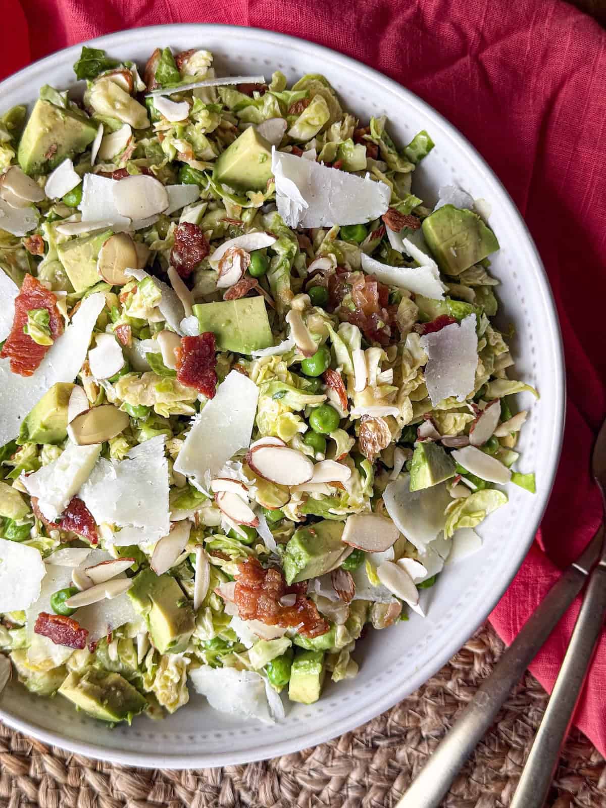 Shredded brussels sprouts salad with peas, chopped bacon, avocado, sliced almonds and shaved cheese in a white bowl with a pink napkin.