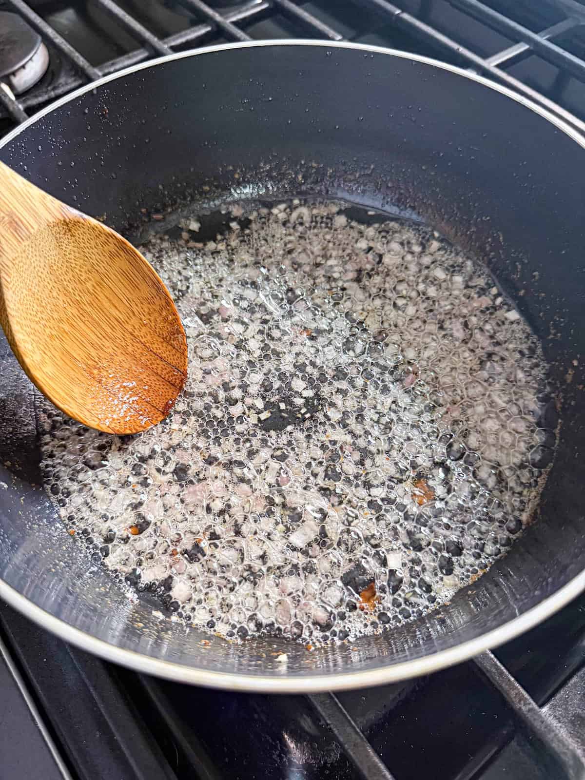 Shallots cooked in bacon grease in a frying pan with a wooden spoon.