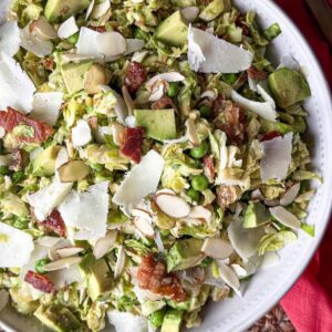 Shredded Brussels sprouts salad with peas, chopped bacon, avocado, sliced almonds and shaved cheese in a white bowl with a pink napkin.