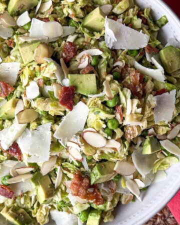 Shredded Brussels sprouts salad with peas, chopped bacon, avocado, sliced almonds and shaved cheese in a white bowl with a pink napkin.