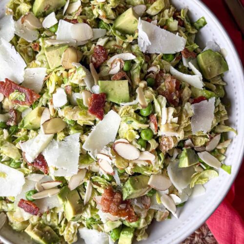Shredded Brussels sprouts salad with peas, chopped bacon, avocado, sliced almonds and shaved cheese in a white bowl with a pink napkin.