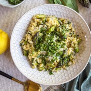 Creamy asparagus and ramp risotto topped with gremolata and Parmesan in a white bowl, served with lemon and a gold spoon.