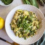 Creamy asparagus and ramp risotto topped with gremolata and Parmesan in a white bowl, served with lemon and a gold spoon.