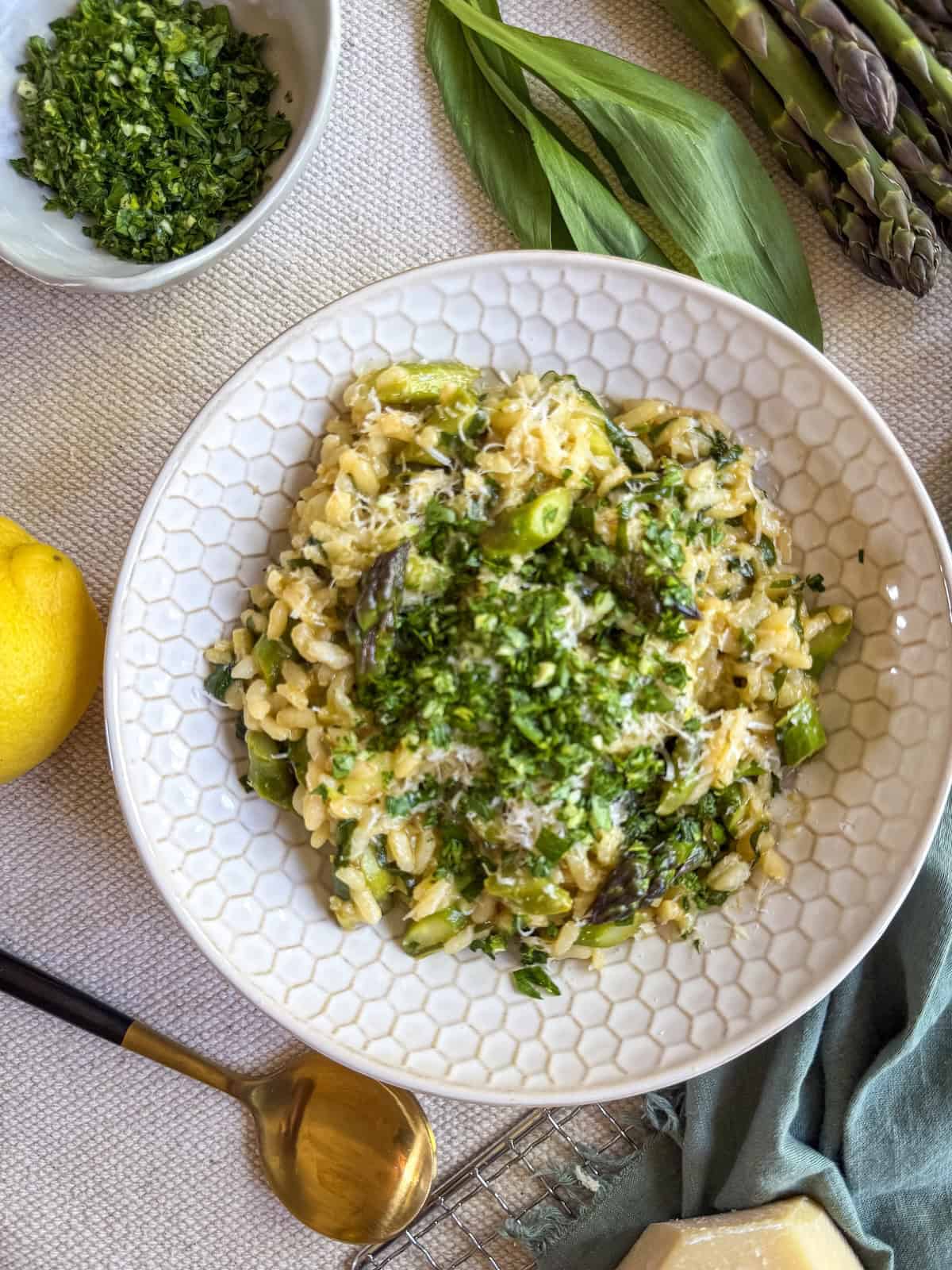 Creamy asparagus and ramp risotto topped with gremolata and Parmesan in a white bowl, served with lemon and a gold spoon.