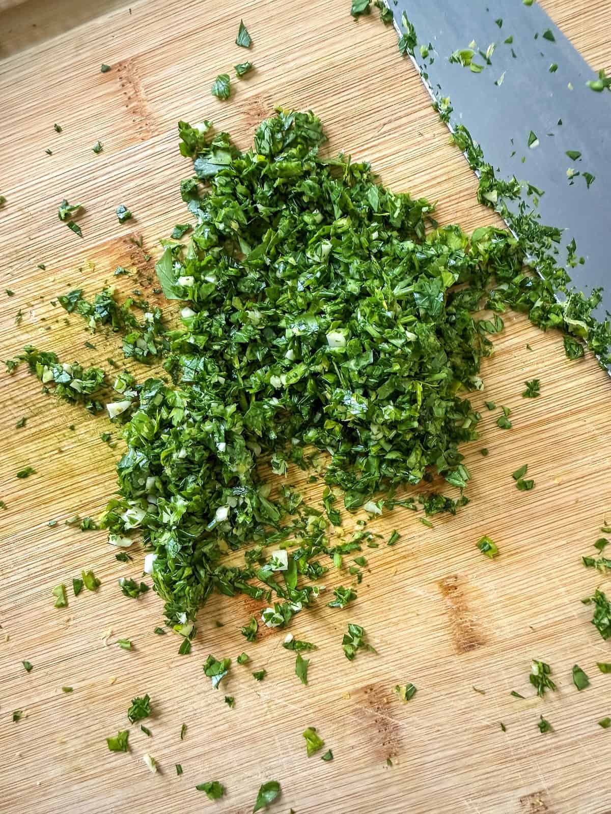 A gremolata of finely minced parsley, garlic and lemon zest on a wood cutting board with a knife.