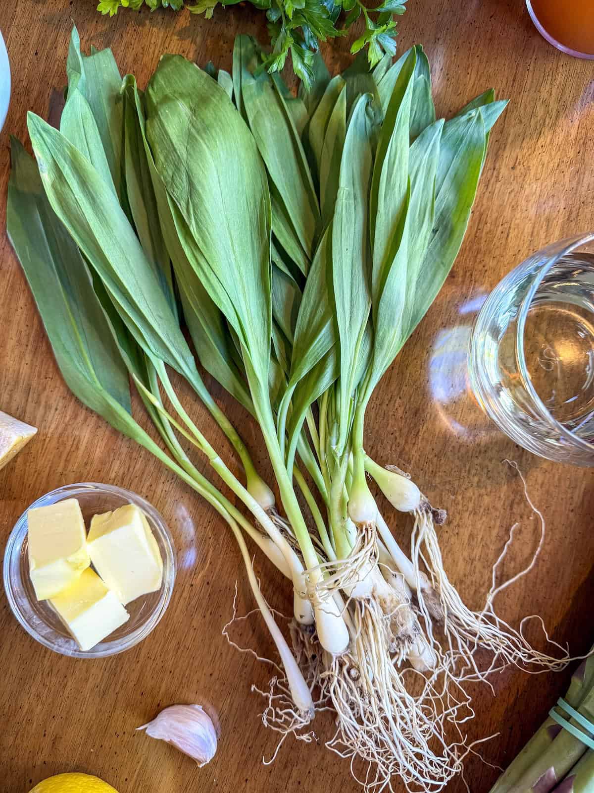 Bunch of ramps on a wood table with a glass of wine and cubed butter in a small glass bowl.