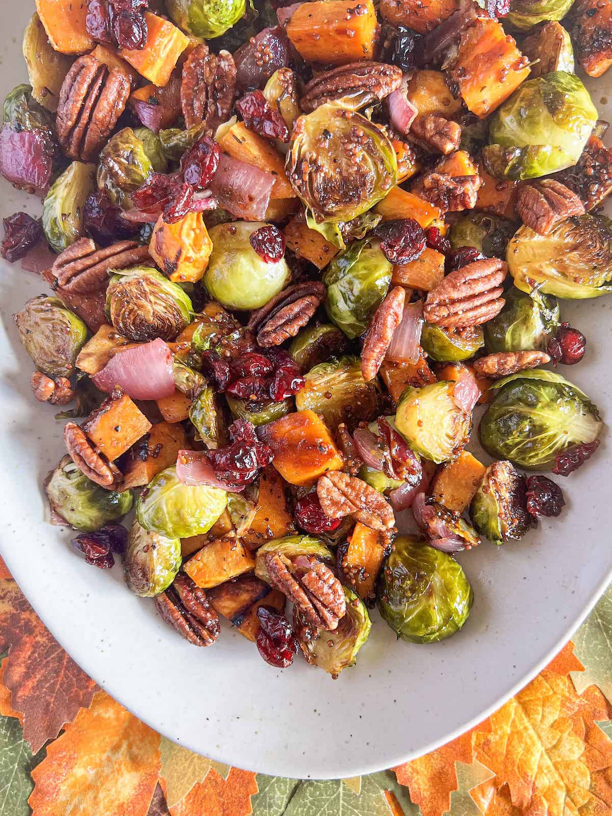 Roasted sweet potatoes and brussel sprouts on a platter topped with candied pecans and dried cranberries.